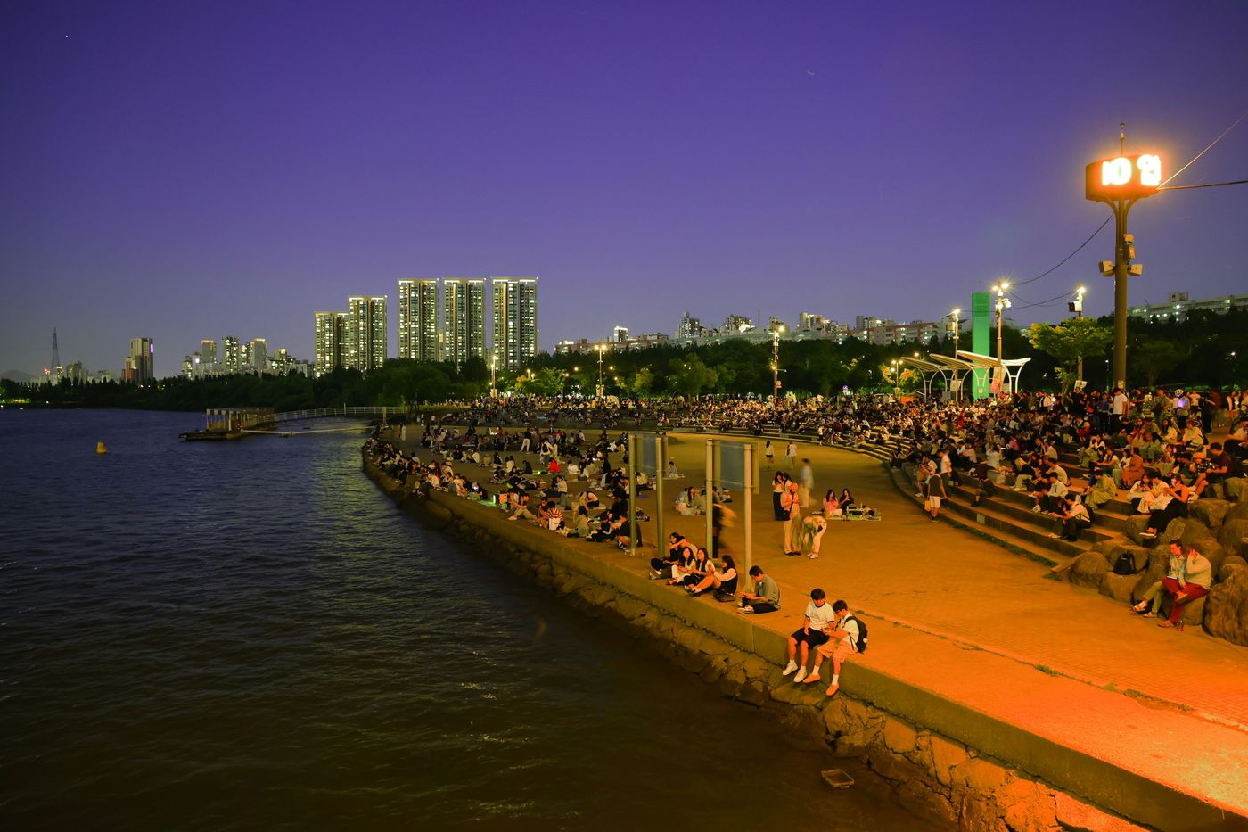 The Han River in Seoul at dusk, people gathered along the water as the city lights come on.